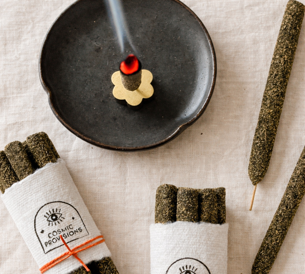 Cosmic Provisions incense bundles on textured linen with an incense stick burning on a black plate and small flower incense holder.