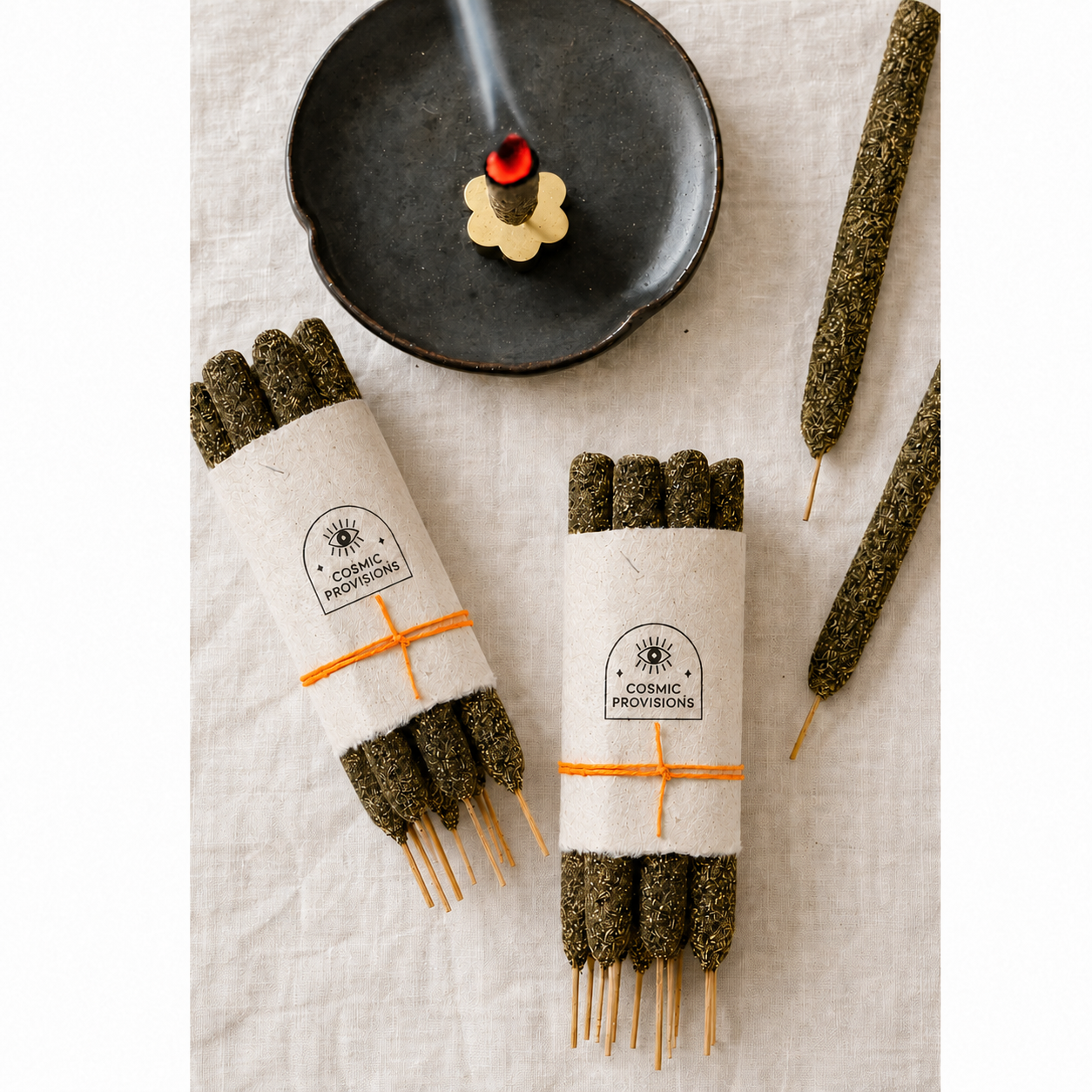 Two bundles of palo + myrrh incense sticks wrapped in lokta paper with orange string and with a 'COSMIC PROVISIONS' logo, arranged on a light linen surface beside two loose incense sticks and a dark ceramic incense burner with  burning incense.