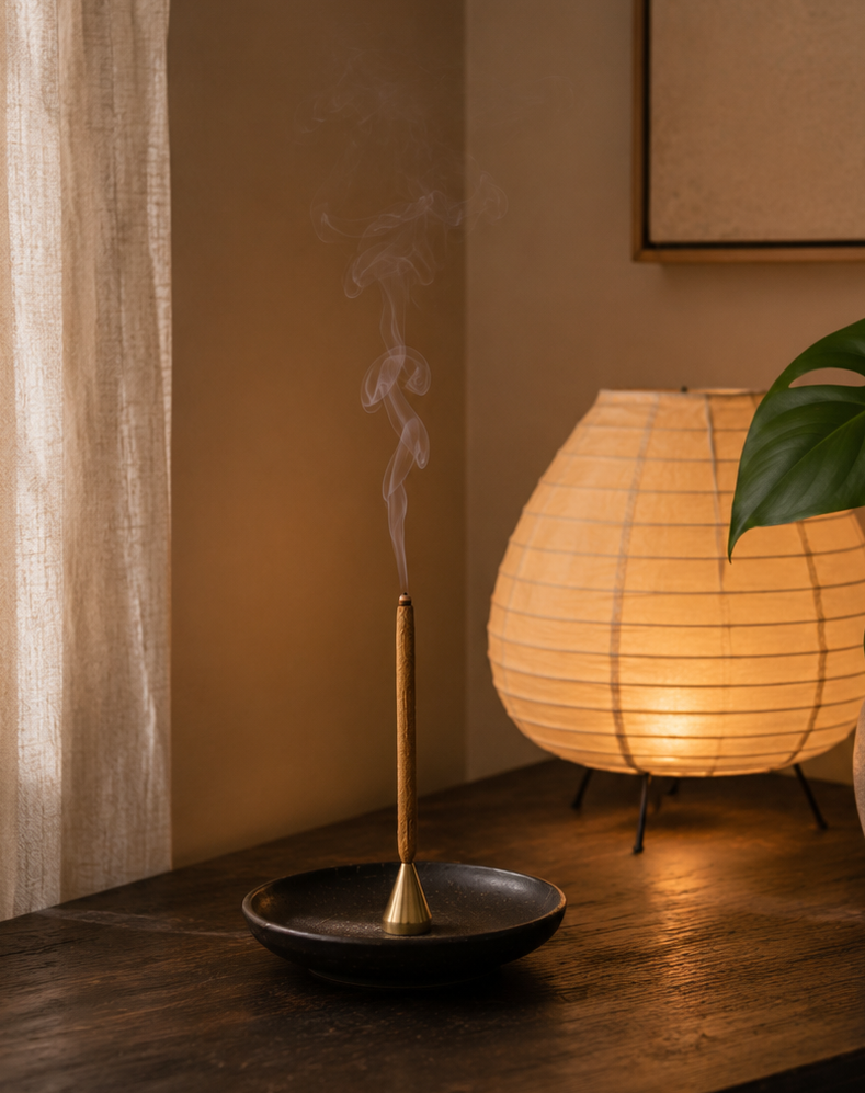 A warm, cozy interior scene featuring a glowing paper lantern table lamp on a dark wooden chest, alongside a brass incense burner with rising smoke and a potted green plant.